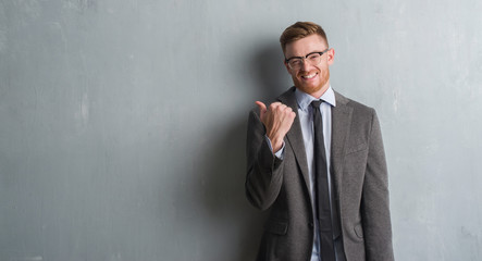 Young redhead elegant business man over grey grunge wall pointing and showing with thumb up to the...