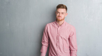 Obraz premium Young redhead man over grey grunge wall wearing pink shirt puffing cheeks with funny face. Mouth inflated with air, crazy expression.