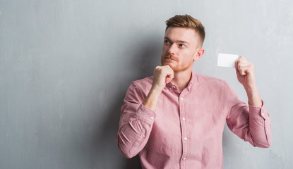 Young redhead man over grey grunge wall holding blank visit card serious face thinking about question, very confused idea - obrazy, fototapety, plakaty