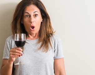 Middle age woman drinking red wine in a glass scared in shock with a surprise face, afraid and...