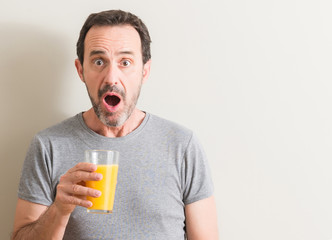 Senior man drinking orange juice in a glass scared in shock with a surprise face, afraid and...