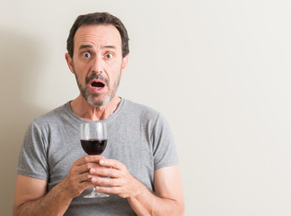 Senior man drinking red wine in a glass scared in shock with a surprise face, afraid and excited...