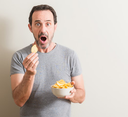 Senior man eating potato chips scared in shock with a surprise face, afraid and excited with fear...
