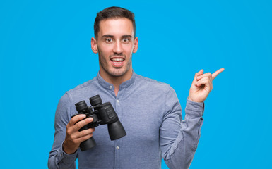 Handsome young man looking through binoculars very happy pointing with hand and finger to the side