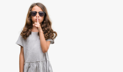 Brunette hispanic girl wearing sunglasses asking to be quiet with finger on lips. Silence and secret concept.