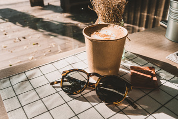 cappuccino in take away coffee cup and sunglasses on table cloth with dry flower on wood table with...