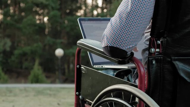 A disabled man is sitting in a wheelchair and working on a laptop in the park