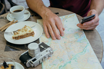 Fototapeta na wymiar A man is holding a smartphone. He is sitting at the cafe table on the street. On the table there is a vintage camera, a tourist map, a coffee and a piece of cake on a plate.