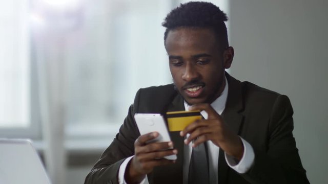 Medium shot of concentrated young black businessman using smartphone to pay online with debit card