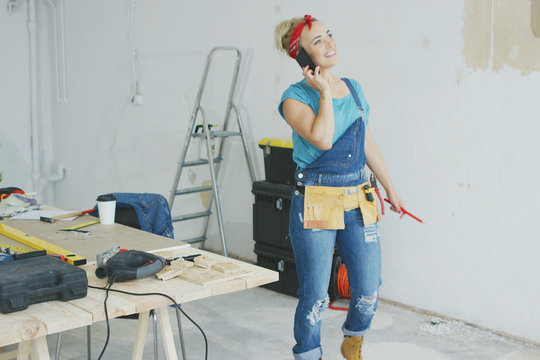Happy smiling young blond woman in jeans overalls standing at carpenter workbench at unpainted wall with stepladder and instruments talking on mobile phone and looking away .