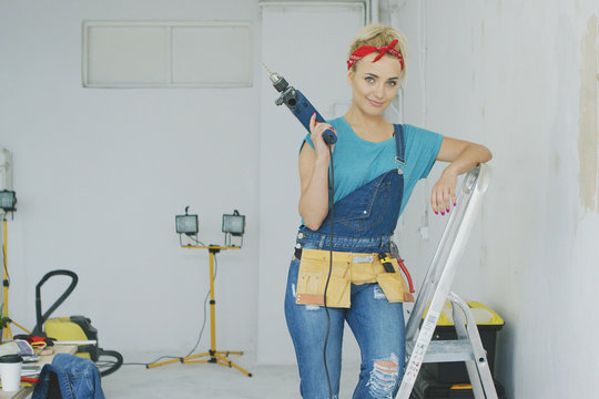 Beautiful blond young female in jeans overalls and red headband standing relaxed leaning on stepladder holding electric drill and looking at camera smiling with unpainted wall and workbench nearby.