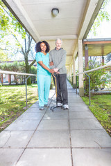 African nurse helping elderly man with crutch in rehab facility hallway