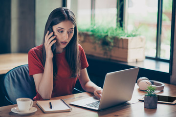 Freelancer business girl in red polo working issues by phone, looks at laptop and fixes errors in documents sitting in cafe