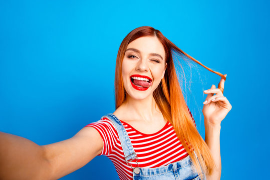 Portrait of nice vivid red cheerful straight-haired excited happy smiling young girl with opened mouth taking self picture, showing tongue out, isolated over blue background