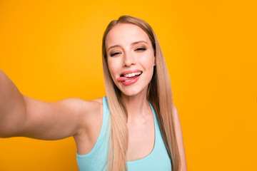 Portrait of happy foolish girl looking camera with tongue out and makes selfie on the front camera of her smartphone isolated on shine yellow background