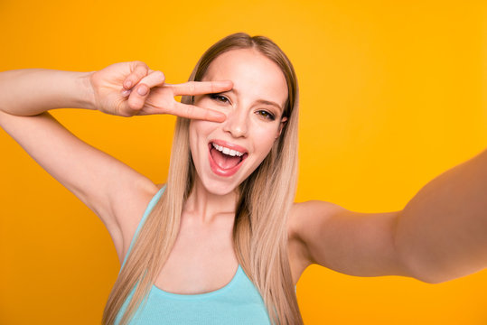 Close up portrait of happy woman with beaming smile making selfie and showing v-sign isolated on shine yellow background