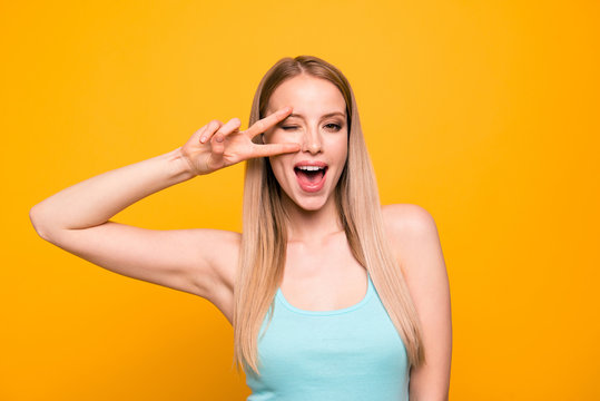 Portrait of foolish cute straight-haired blonde caucasian girl, wearing casual blue shirt, showing v-sign. Isolated over yellow background
