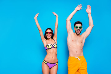 Hot summer time concept. Portrait of cheerful couple in sunglasses and bright swimsuits enjoying sunlight together with beach party mood raise their hands up isolated on bright blue background