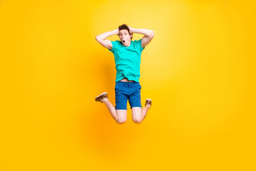 Fototapeta na wymiar Young attractive handsome funky curly-haired excited guy wearing casual green polo, shorts, sneakers, jumping up in the air, holding hands on head. Isolated over vivid shine bright yellow background