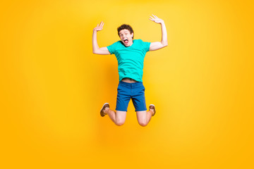Fototapeta na wymiar Young attractive handsome curly-haired excited guy wearing casual green polo, shorts, sneakers, jumping up in the air, raising hands up, joyful. Isolated over vivid shine bright yellow background