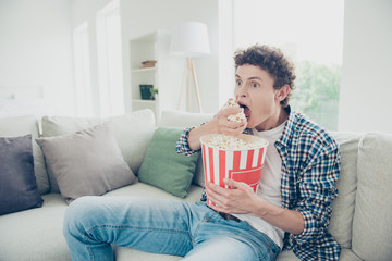 Fototapeta premium Attractive curly-haired concentrated young guy student wearing casual, sitting of sofa at home, grabbing eating pop corn, watching tv reality show