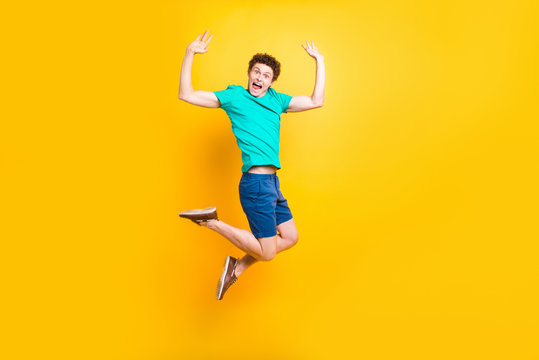 Full size length body picture of handsome curly-haired playful young guy wearing casual green t-shirt, shorts, shoes, jumping in air, raising hands up. Isolated over yellow background