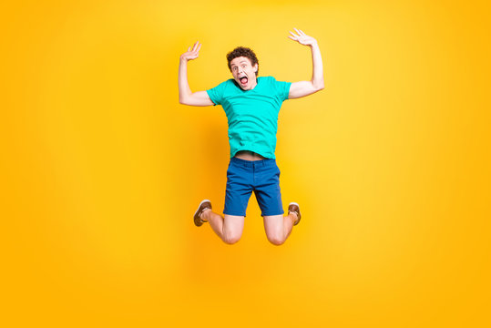 Young attractive handsome curly-haired excited guy wearing casual green polo, shorts, sneakers, jumping up in the air, raising hands up, joyful. Isolated over vivid shine bright yellow background