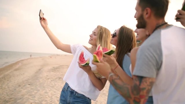 Friends Eating Watermelon Standing on Sandy Beach and Taking Selfies with Smartphone Camera. Young Men and Women Wearing Blue Jeans Shorts Near Sea ans Talking. Healthy Nutrition, Vegatarian Food