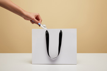 cropped image of woman putting paper hat into shopping bag isolated on beige