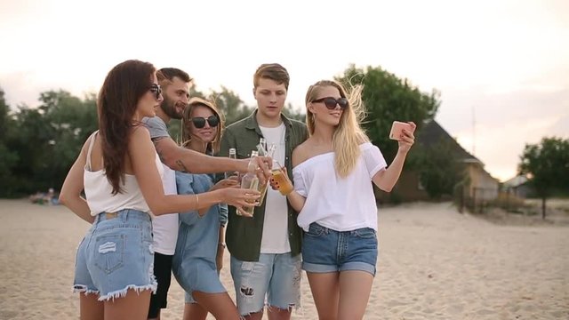 Group of friends having fun enjoying a beverage and relaxing on the beach at sunset in slow motion. Young men and women drink beer standing on a sand in the warm summer evening.