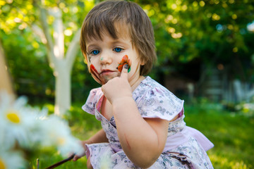little girl is drawing. Girl learns to paint. Young artist. child dirty his face with paints. Funny girl is engaged in creativity in the park. 
