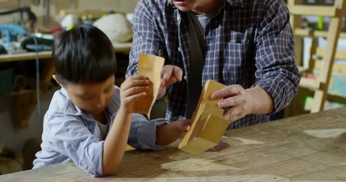 Handheld shot of cute Asian boy sanding wooden toy in carpentry workshop as his father giving him advice