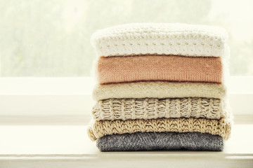 Bunch of knitted warm pastel color sweaters, different knitting patterns folded in stack on white windowsill, window view background. Fall winter season fashionable knitwear. Close up, copy space.