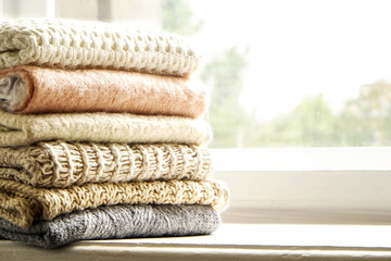 Bunch of knitted warm pastel color sweaters, different knitting patterns folded in stack on white windowsill, window view background. Fall winter season fashionable knitwear. Close up, copy space.