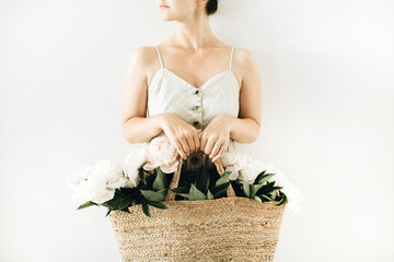Young pretty woman holding straw bag with white peony flowers on white background. Summer concept.