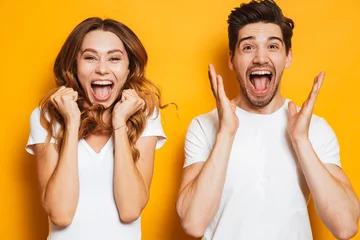 Deurstickers Photo of beautiful admired couple man and woman in basic clothing screaming in surprise or delight and touching cheeks, isolated over yellow background © Drobot Dean