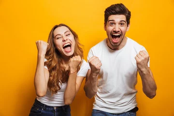 Foto op Canvas Portrait of positive beautiful man and woman in basic clothing screaming and clenching fists like winners or happy people, isolated over yellow background © Drobot Dean