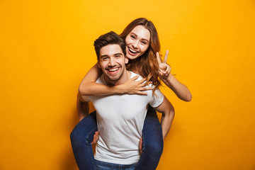Image of caucasian couple having fun while man piggybacking joyful woman, isolated over yellow background - obrazy, fototapety, plakaty