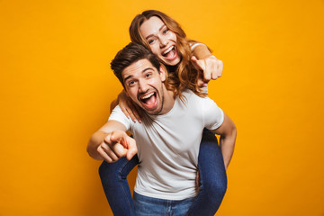 Image of caucasian man giving piggyback ride to joyful woman and pointing fingers at you, isolated over yellow background