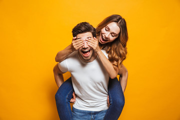 Image of loving couple having fun while man piggybacking joyful woman, isolated over yellow background