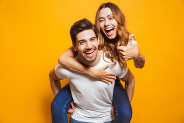 Image of excited couple having fun while man piggybacking joyful woman with thumb up, isolated over yellow background