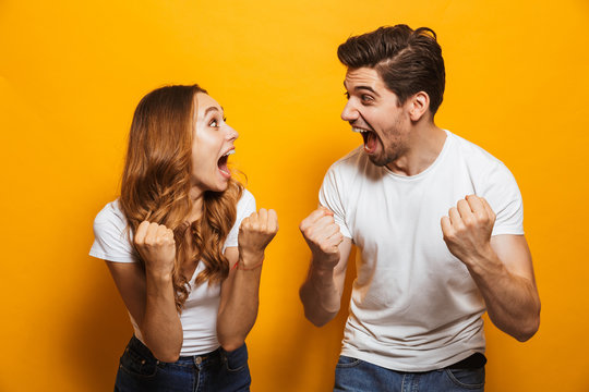 Photo of delighted european man and woman in basic clothing screaming and clenching fists like winners or happy people, isolated over yellow background
