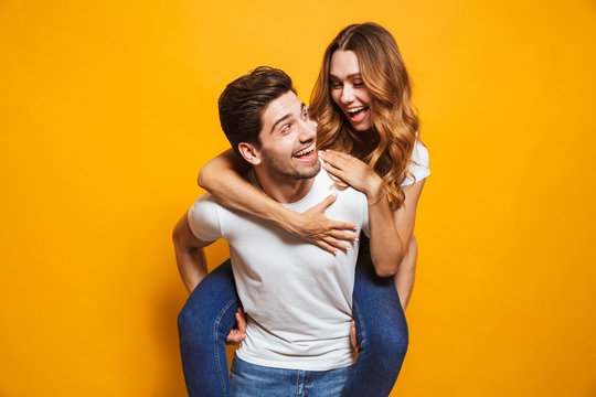 Image of caucasian couple having fun while man carrying beautiful woman on his back, isolated over yellow background