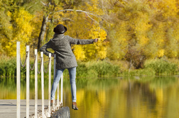 Girl in a hat with leaves in hands standing on the dock. Autumn, sunny. Back view