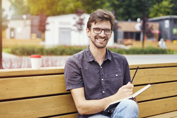 Handsome young man smiling face writing in a note book sitting outside. Guy wearing glasses alone working. Concept of education students youth.