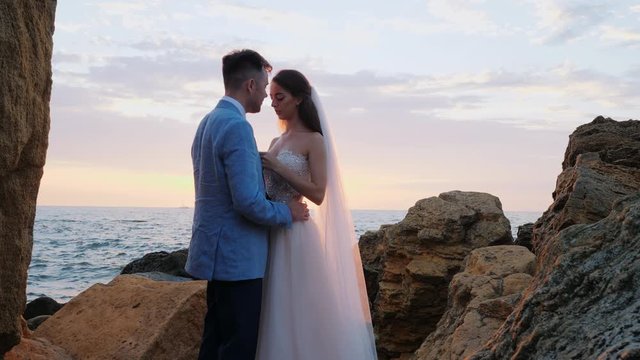 Beautiful young wedding couple standing on sea shore with rocks. Newlyweds spend time together: embrace, kiss and care for each other. Love concept. Bride smiling to groom.