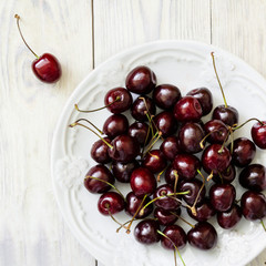 Dark sweet cherry in a white plate and on light wooden table close-up. Top view.