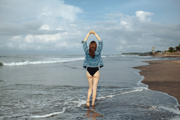 Fototapeta na wymiar Happy smile brunette Woman wearing black swimsuit and denim jacket at ocean background enjoy walking at beach, hands raised up to the sky. Freedom concept