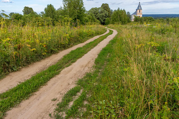Fototapeta na wymiar Vanishing dirt road to church through meadow overgrown by cow-parsnip. Berezichi, Kaluzhsky region, Russia