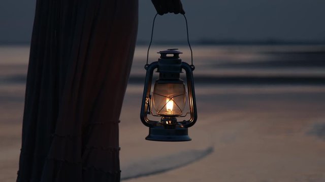 Girl in dress holding old kerosene lantern in her hand on beach after sunset at night